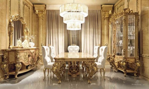 This picture shows a luxurious dining room interior designed with a modern flare. The walls are painted a deep green color, and the ceiling is adorned with intricate gold and white moulding details. The furniture is mostly composed of white seating and dining pieces, accented with golden detailing and glass accents such as the coffee table. A long white buffet table on one side offers storage space, while a statement chandelier hangs from the middle of the room, giving off a warm glow of light.
