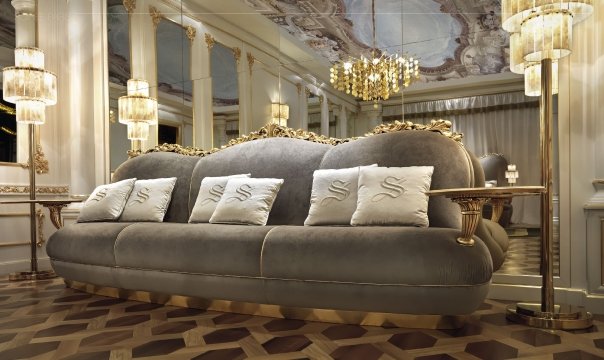 This picture shows a luxurious living room that has been professionally decorated. It has a classic, traditional style with touches of modern elements. There are white walls, a marble fireplace, intricate moldings, and a grand chandelier in the center of the room. The room is filled with dark wooden furniture including an armchair, chaise lounge, and side tables. The sofa has a bright patterned fabric, which adds a touch of color to the otherwise neutral room. There is a large area rug in the center of the room and a selection of artwork on the walls.