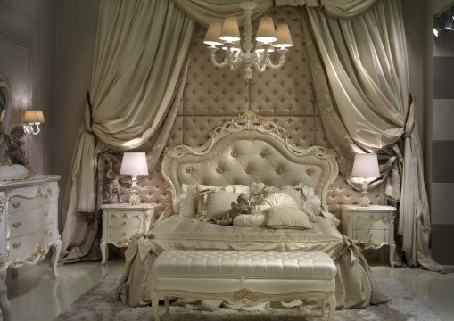 This picture shows an elegant and luxurious living room, with sophisticated furniture and décor. The walls have been painted a soft cream color, and the floor is covered in a plush beige carpet. The sofa is a white leather tufted design with gold accents. There is a large crystal chandelier hanging from the ceiling, adding a regal touch to the room. In front of the sofa is a white marble coffee table and two ornately designed side tables. On the walls are a few tasteful paintings and golden framed mirrors. The overall design of the living room makes it the