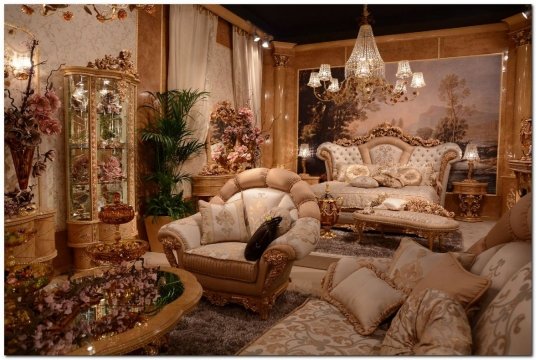 This picture shows a luxury home interior with a large, ornate, light-colored sofa in the center of the room. The sofa is surrounded by various high-end furniture pieces and accent pieces, including a round coffee table, a rug, a couple of delicate side chairs, and an intricate chandelier. There are elegant drapes decorating the floor-to-ceiling windows and two beautiful abstract paintings on the walls. Everything in the room looks very expensive and gives off an upscale vibe.