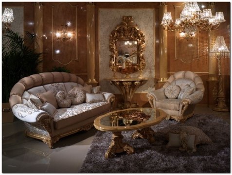The picture appears to be a modern, luxury living room. The walls are cream-colored and there is an ornate white stone fireplace on the left side of the room. There are two dark brown leather couches in the center of the room facing each other, accompanied by two glass-top tables. There is a beige area rug underneath the seating area and a large black chandelier hanging from the ceiling. The walls are adorned with various pieces of art, such as a painting above the fireplace and a series of sculptures. Further to the right, there is a grand piano and