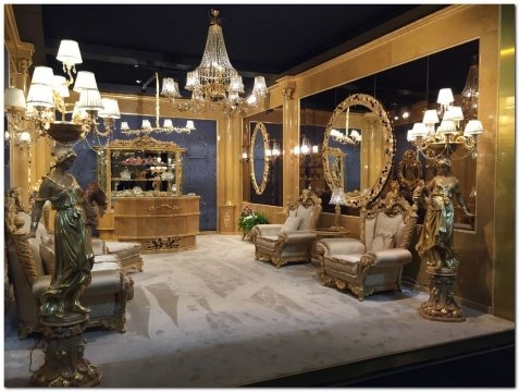 This picture shows a luxurious living room with an impressive marble wall and floor. There is a large beige sofa in the middle of the room with accent cushions and a white fur rug in front of it. The walls are decorated with framed artworks and there is gold-framed mirror on the wall. A modern chandelier hangs from the ceiling, providing dramatic lighting to the room.