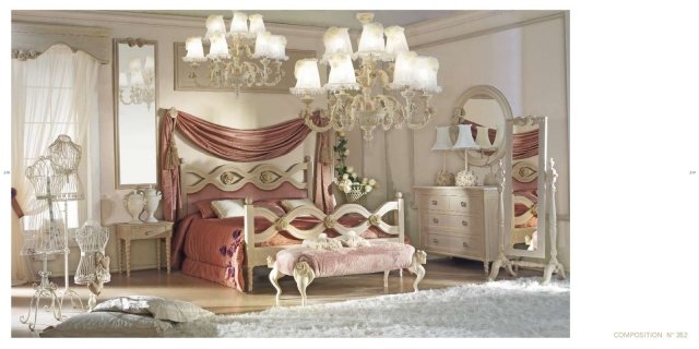 This picture shows a modern luxury bedroom design, which features a large upholstered bed with white and cream pillows. There is a statement headboard with a mirror above the bed and two side tables with lamps on each side of the bed. The room also has a grey chaise lounge, a dark fireplace and ornate wall art on the walls. The floor is laid with a light beige carpet and there are draperies hung in the corners of the room.