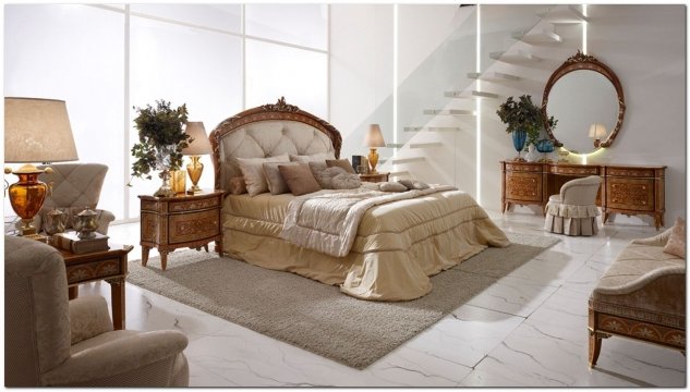 The picture shows a beautiful modern bedroom with a four-poster bed, two nightstands, and a grey and brown dresser. The walls are painted a light beige color and there is a large white window. There is a patterned area rug in front of the bed, and several pieces of contemporary furniture in the room. A floor lamp stands in one corner and several framed pieces of art hang on the wall adding to the decor.