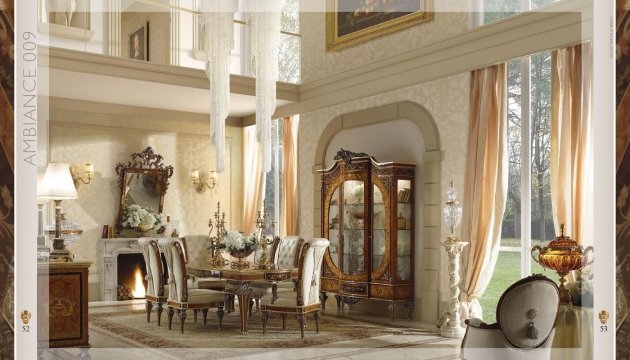 The picture shows a luxurious home interior. It features crystal chandeliers and light fixtures, an ornate marble staircase with a gold railing, white marble walls and flooring, and plush beige sofas with matching chairs. The walls are decorated with framed artwork and mirrors. The ceiling has subtle decorative details and a large crystal chandelier.