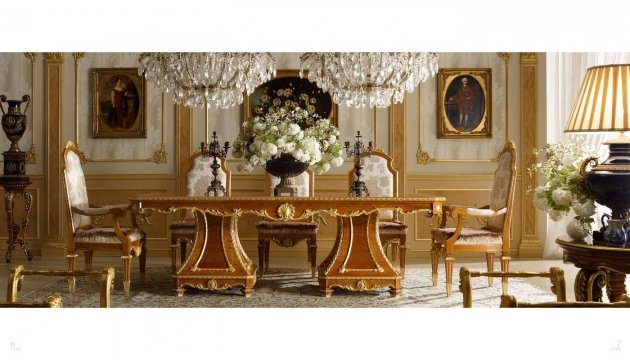 This picture shows an elegant living room with a white and gold decor. There is a luxurious golden sofa in the center of the room, with two matching armchairs on either side. A glass coffee table with a decorative flower arrangement sits in the middle, and a two-tier side table with a lamp and houseplant stands at the side. There is an elaborate ceiling light and two wall sconces on either side of the room. The walls are painted a pale cream color, while the floor is covered in a plush beige rug.