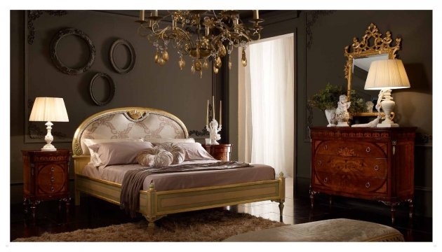 The picture shows an elegant and luxurious master bedroom. The room features an elegant tufted headboard in a rich golden color, a matching footboard with gold accents, and matching night stands. The walls are painted in a warm beige color, while the floor is covered in a unique patterned white and beige marble tile. In the corner is a plush velvet armchair with a footstool in front of it. An ornate gold and crystal chandelier hangs from the ceiling, adding a touch of sophistication to the room.