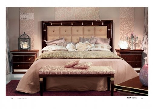 This picture shows a classic and luxurious bedroom design. The room features a high ceiling with detailed crown molding and wood paneling along the walls. A large, tufted headboard is situated against the wall, providing a comfortable place to rest. The bed is topped with a classic floral comforter set, which is surrounded by several pillows. The bed is flanked by two matching nightstands that feature intricate designs and crystal knobs. The floor is covered with a plush white rug and several armchairs are situated at the foot of the bed. A crystal chandelier hangs